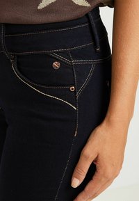 Mörka jeanstyg med kontrasterande beige sömmar. Funktioner inkluderar en framficka med en liten metallaccent och en figurnära passform.