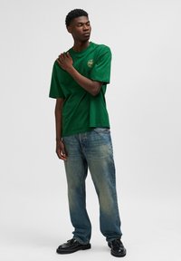 Grön bomullströja med avslappnad passform, rund hals, och en liten grafisk logotyp på bröstet, i par med urtvättade blå jeans och svarta skor.