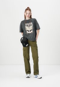 Schwarzes Grafik-T-Shirt mit oversized Passform und Schmetterlingsmotiv. Olivgrüne Cargo-Hose mit Seitentaschen. Weiße Sportschuhe, schwarze Kappe.