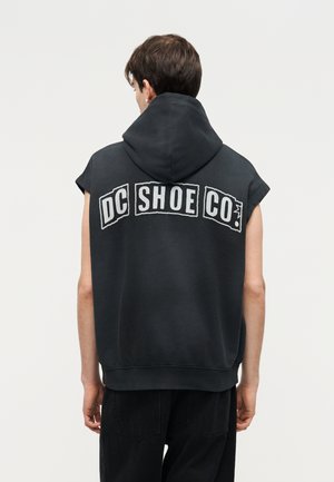 Νεαρό άτομο φαίνεται από πίσω, φορώντας μαύρο αμάνικο φούτερ με κουκούλα με το λογότυπο «DC SHOE CO.» στην πλάτη και μαύρο παντελόνι.