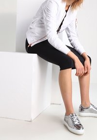 Witte bomberjas met geplooide details, gecombineerd met zwarte leggings tot op de knie en metallic zilveren sneakers. Overal een gladde textuur.