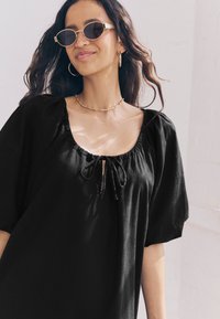 Sort hørbluse med afslappet pasform, puffede korte ærmer, indsamlet halsudskæring og justerbar bindedetalje. Model iført guldtilbehør.