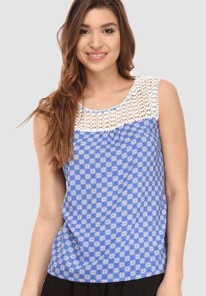 Joven mujer con cabello largo castaño sonríe, vistiendo una blusa sin mangas de color azul con un patrón y un escote de ganchillo blanco.