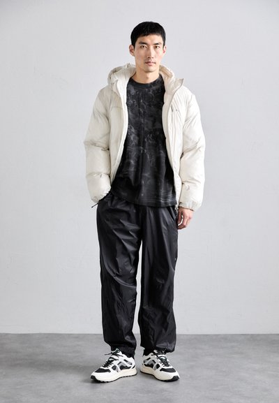 Chaqueta tipo plumón beige claro con capucha, camiseta negra con estampado, pantalones impermeables negros y zapatillas blancas con acentos negros. Diseño minimalista.