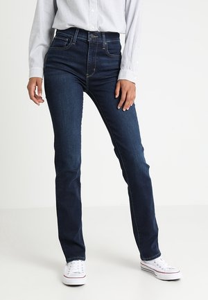 Osoba ubrana w obcisłe ciemnoniebieskie dżinsy, białe trampki oraz jasnoniebieską koszulę w paski, stojąca na tle jednolitej białej ściany.