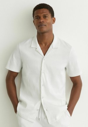 Jonge man die een wit gestructureerd kortermig overhemd met kraag draagt, handen in de zakken, staand tegen een witte muur.