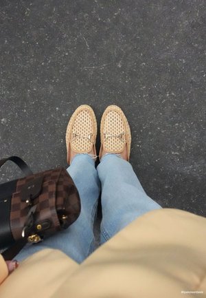 Mocassins tissés beiges portés avec un jean bleu clair, debout sur un sol texturé sombre, tenant un sac à main marron à carreaux.