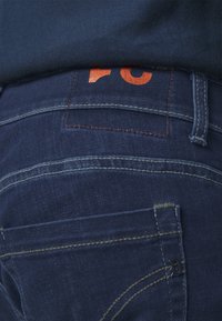 Primer plano de un bolsillo trasero de unos vaqueros de mezclilla azul oscuro con una etiqueta de marca cosida en naranja parcialmente visible por encima de la cintura.