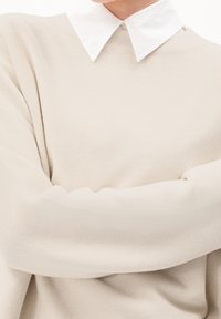 Beżowy sweter o fakturowanym splocie i podwiniętych mankietach, noszony na białej koszuli z kołnierzykiem. Charakteryzuje się luźnym krojem i miękkim materiałem.