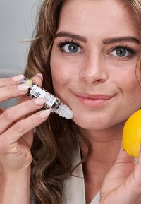 Bouteille transparente de rollerball d'huile avec une étiquette noir et blanc, tenue près d'un visage, accompagnée d'un citron jaune, sur un fond neutre.