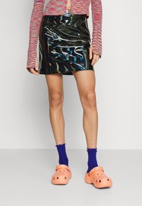 Mini-jupe en vinyle noir brillant avec une texture lisse, associée à des chaussettes côtelées violettes éclatantes et des sabots orange.