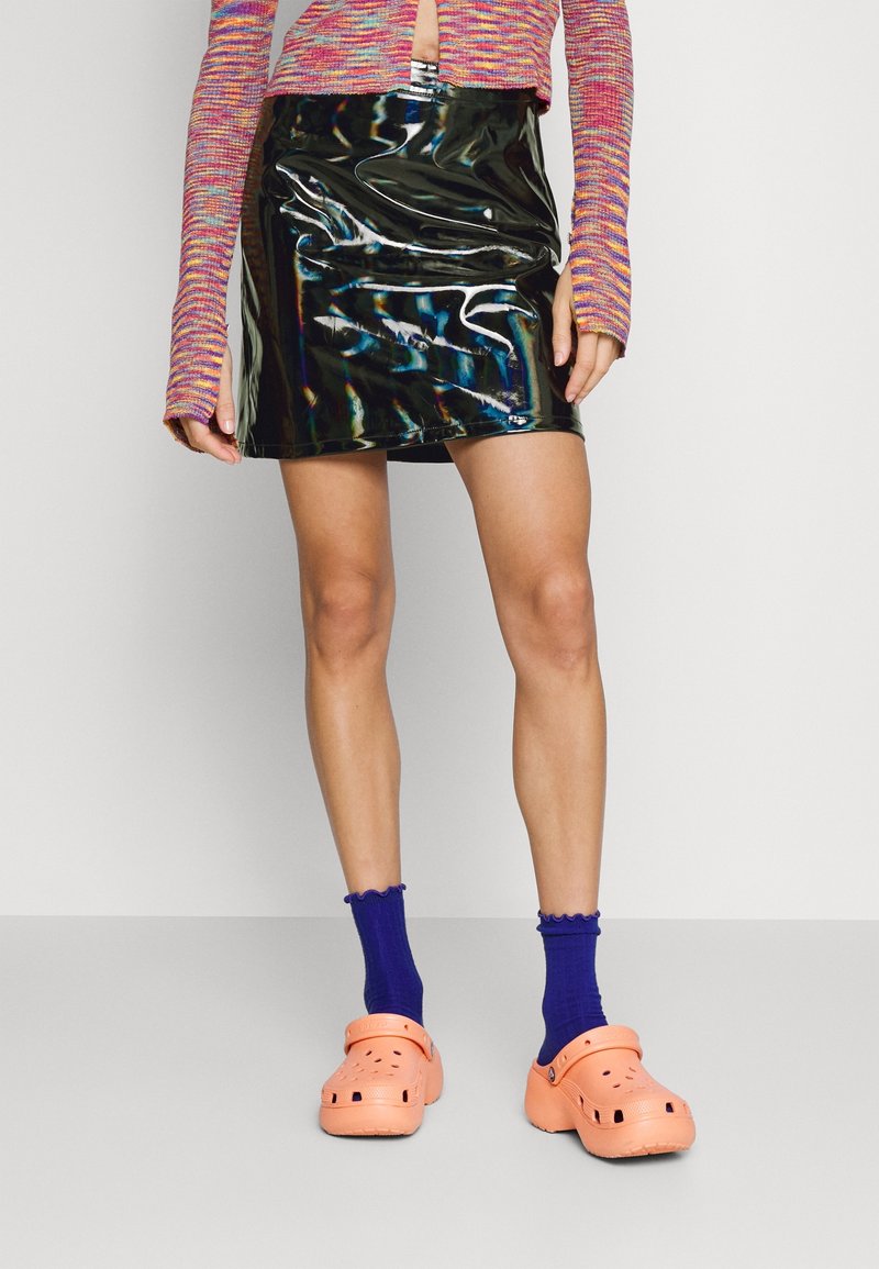 Mini-jupe en vinyle noir brillant avec une texture lisse, associée à des chaussettes côtelées violettes éclatantes et des sabots orange.