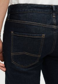 Κοντινή λήψη πίσω τσέπης και ζώνης σε σκουρόχρωμα μπλε τζιν, φορεμένα με λευκό πουκάμισο, εμφανίζοντας αντίθετη ραφή και θηλιές για ζώνη.