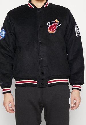 Osoba nosząca czarną kurtkę varsity Miami Heat z naszywkami z logo NBA, czerwono-białymi paskami na mankietach i zapięciem na guziki.