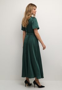 Rochie maxi din satin verde cu mâneci scurte, talie ajustată și fustă fluidă, asortată cu tocuri stiletto negre. Vedere din spate, fundal simplu.