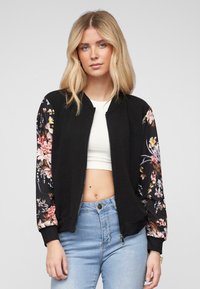 Schwarze Bomberjacke mit floral gemusterten Ärmeln; verfügt über gerippte Bündchen und einen Reißverschluss. Getragen über einem weißen Cropped-Top und blauen Jeans.