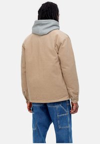 Chaqueta de pana marrón con capucha gris, que presenta una forma relajada, textura acanalada y puños abotonados. Pantalones de mezclilla con múltiples bolsillos.