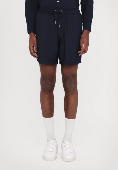Shorts bleu marine avec une taille élastique et un cordon de serrage, présentant une texture lisse et une coupe décontractée, associés à des baskets blanches et des chaussettes.