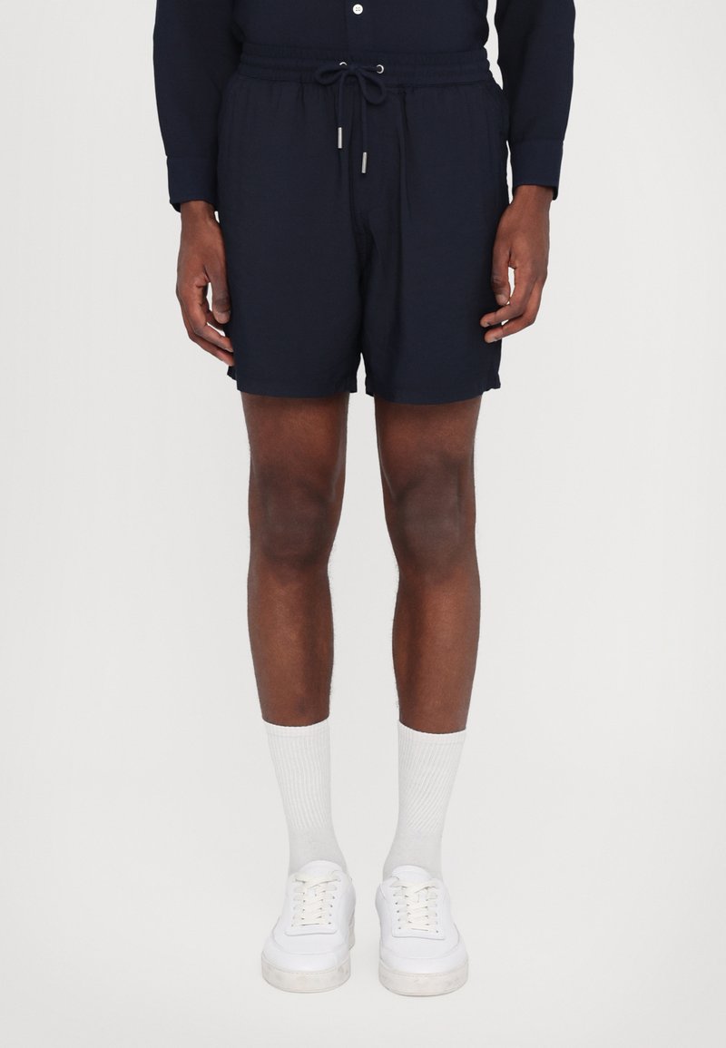 Mørkeblå shorts med elastisk talje og snor, som har en glat tekstur og en afslappet pasform, parret med hvide sneakers og sokker.