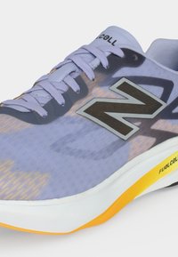 Ανοιχτόμωβ αθλητικό παπούτσι New Balance με μαύρο λογότυπο "N", λευκή και κίτρινη σόλα και το κείμενο "FuelCell" στο πλάι κοντά στη φτέρνα.