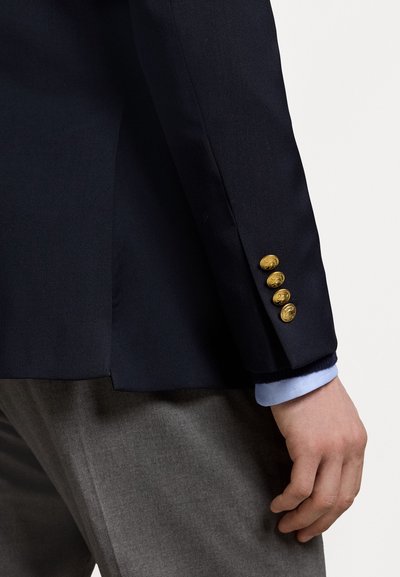 Marineblazer met vier gouden knopen op de mouwen, gecombineerd met een lichtblauw shirt en grijze broek. Gladde stof, gestructureerd ontwerp.