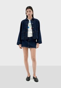 Denim jacket with a relaxed fit, button closure, two front pockets, paired with a white graphic t-shirt and a dark denim skirt, worn with black shoes.