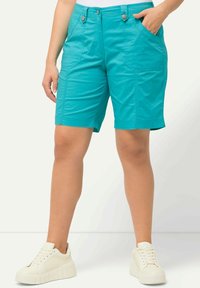 Pantalones cortos turquesa con una textura suave, que cuentan con dos bolsillos delanteros, detalles de botones y un diseño a la altura de la rodilla. Combinados con zapatillas blancas.