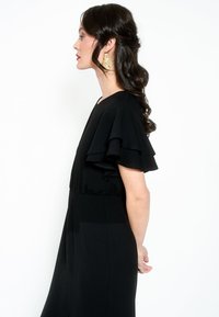 Vestido negro con mangas acampanadas y en capas, cintura ajustada y textura suave. Pendientes de hoja dorada añaden interés visual. Modelo mostrada de perfil.