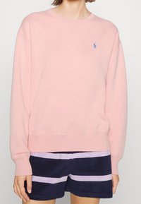 Sweatshirt rose clair en coton doux, avec un col rond et des poignets côtelés. Un petit logo bleu brodé est présent sur la poitrine.