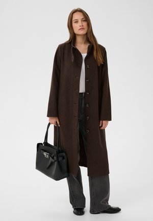 Femme debout face à l’avant, portant un long manteau marron à boutons, un haut blanc, un pantalon gris, des chaussures noires et tenant un grand sac à main noir.