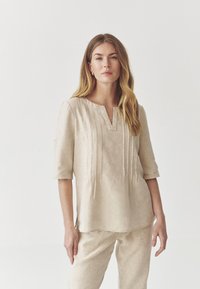 Beige lněná blůza se třičtvrtečními rukávy, výstřihem do V a vertikálními sklady vpředu. Nohavice jsou sladěny s lněnými kalhotami.