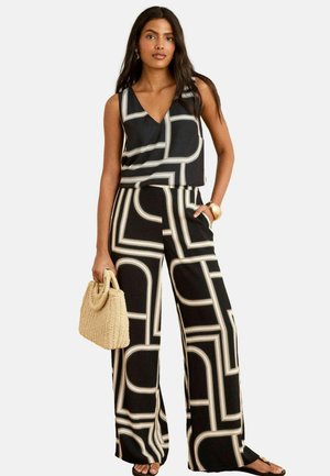 Schwarz-weiß gemusterter Jumpsuit mit V-Ausschnitt und weitem Bein, der geometrische Designs aufweist. Das Model hält eine gewebte beigefarbene Tasche.