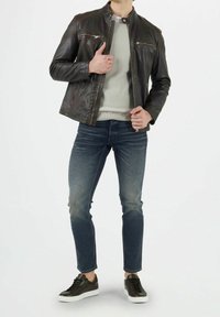 Brązowa skórzana kurtka z kołnierzem stójkowym, dwoma kieszeniami na zamek na piersiach, noszona na jasnoszarym swetrze, w połączeniu z dopasowanymi niebieskimi dżinsami i czarnymi sneakerami.