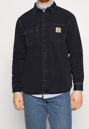 Mand iført sort knap-op jakke over lyseblå skjorte og blå jeans, med to brystlommer og synligt Carhartt-logo på venstre lomme.