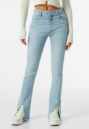 Jeans acampanados de color azul claro con aberturas laterales en el dobladillo, confeccionados en denim. Presentan una cintura ajustada y un diseño de cinco bolsillos, combinados con zapatillas blancas.
