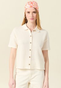 Kortärmad, krämfärgad polo med ett strukturerat mönster, knäppning framtill och en kontrasterande krage. Bärs med ljusa byxor och en blommig huvudduk.