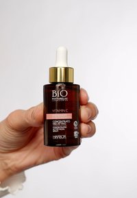 Brun glasflaska med ett guldlock, pipettopp och etiketter i vitt och rosa. Innehåller texten: "Vitamin C," "Koncentrerat aktivt ansiktsserum."