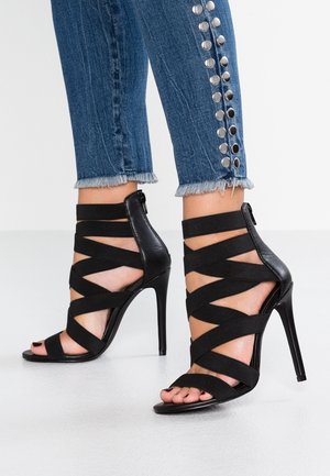 Sandales à talons noirs avec des brides élastiques croisées, bout ouvert et fermeture éclair à l'arrière. Associées à un jean court en denim orné de clous métalliques.