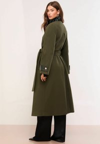 Płaszcz w kolorze oliwkowej zieleni z paskiem w talii, kołnierzem oraz błyszczącymi czarnymi akcentami na kołnierzu i mankietach. Długa, prosta sylwetka, dopasowany krój.