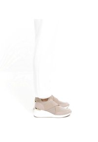 Baskets de sport beiges avec des sangles élastiques croisées, des panneaux en maille et des détails texturés. Semelle blanche avec un léger talon compensé et des accents métalliques.