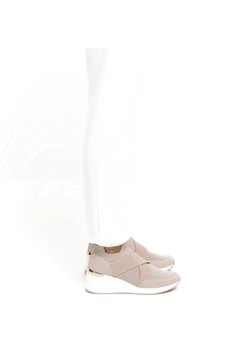 Baskets de sport beiges avec des sangles élastiques croisées, des panneaux en maille et des détails texturés. Semelle blanche avec un léger talon compensé et des accents métalliques.