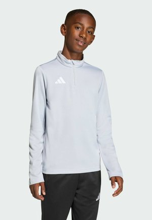 Teenage dreng iført en lysegrå sports-top med kvart-lynlås og sorte Adidas bukser, stående mod en ensfarvet lys baggrund og smilende let.