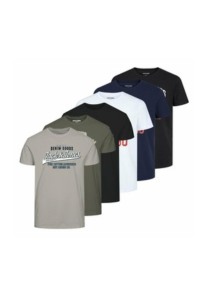 Mężczyzna ubrany w białą graficzną koszulkę Jack & Jones, ciemnoniebieskie jeansy i białe trampki, stojący ze splecionymi rękami na szarym tle.