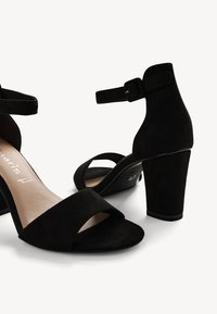 Black suede high-heeled sandals with an ankle strap and open toe design. Features a wide front band and a chunky, textured heel.