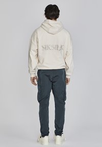 Cremefarbener Hoodie mit "SKSILK"-Logo auf dem Rücken, kombiniert mit dunkelblauen Cargo-Hosen und weißen Sneakers. Lässiger Schnitt, weicher Stoff und verstellbare Kapuze.