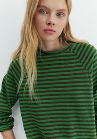 Giovane donna con lunghi capelli biondi e occhi azzurri, che indossa una maglia a maniche lunghe a righe verdi e marrone scuro e un orecchino a cerchio d'argento.