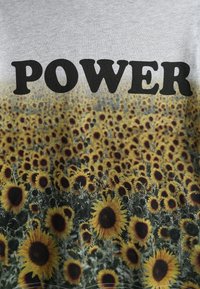 Harmaan T-paita, jossa lukee sana "VOIMA" paksulla mustalla fontilla värikkään auringonkuntakentän päällä, jossa on keltaisia terälehtiä ja tummia keskikohtia.