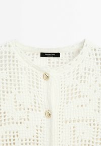 Hvid strikket cardigan med åbent vævedesign, rund halsudskæring og lukning foran med guldknap. Mærkeetiket synlig.