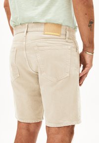 Beige katoenen shorts met een rechte snit, zichtbare achterzakken en een tan leren patch met de tekst "ARMEDANGELS" op de tailleband.