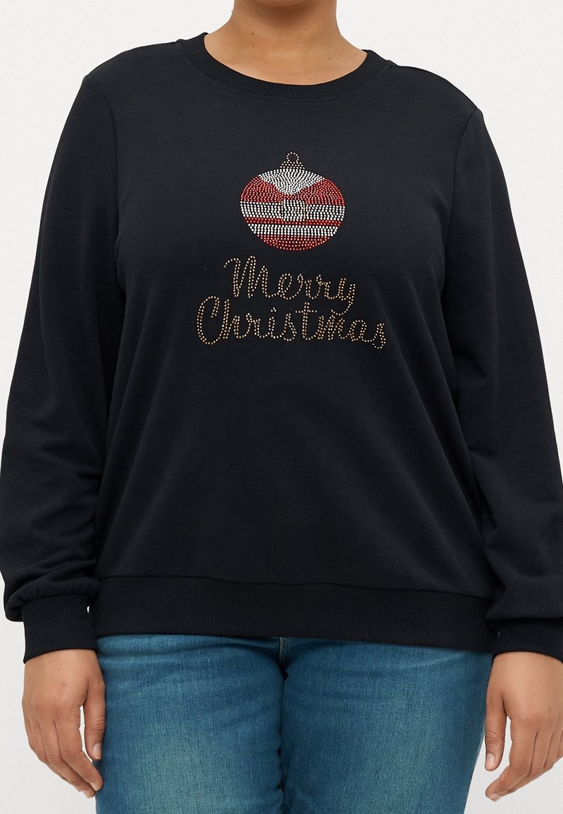 Pull noir à manches longues avec motif ornemental rouge et blanc et texte « Joyeux Noël » en clous dorés sur le devant, porté avec un jean bleu.